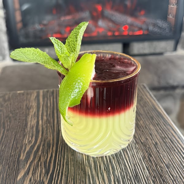 A layered cocktail with yellow and red liquids, garnished with mint and lime, in front of a fireplace.